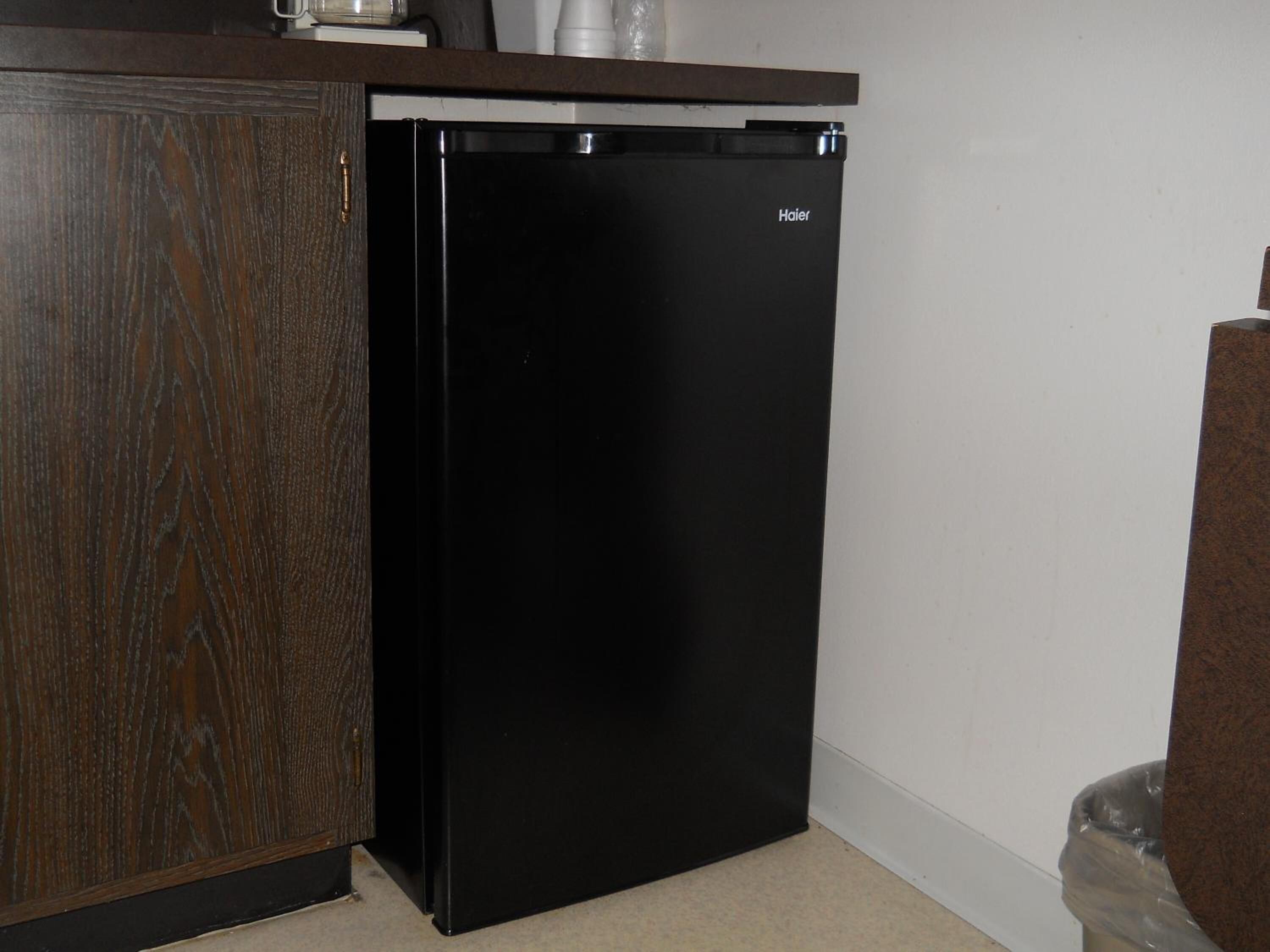 Un refrigerador negro compacto con el logotipo de Haier, ubicado frente a una pared blanca en una cocina con un armario de madera y un bote de basura.