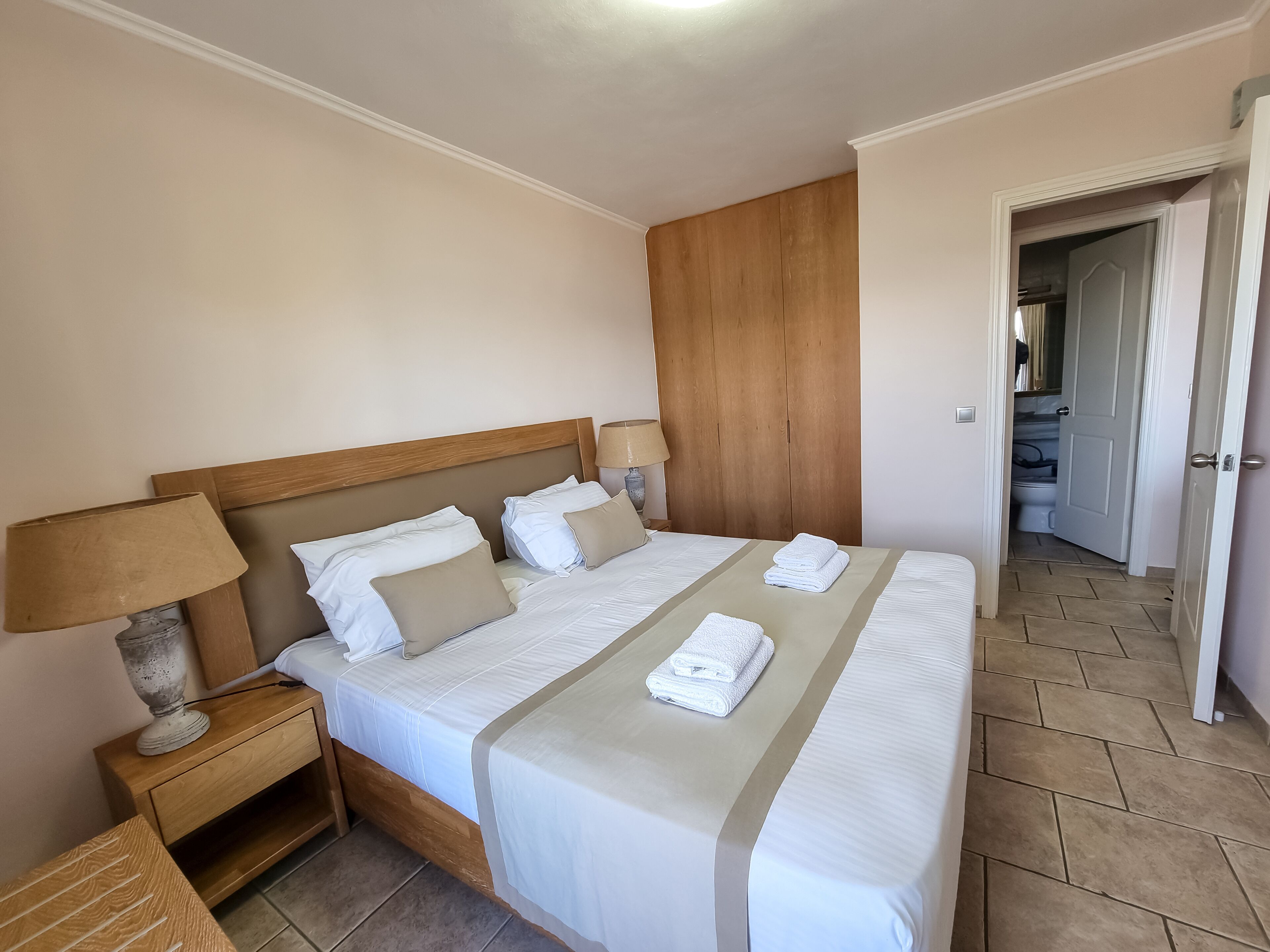 A neatly made double bed with white linens and beige pillows, a wooden headboard, a bedside table with a lamp, and a wardrobe in the background.