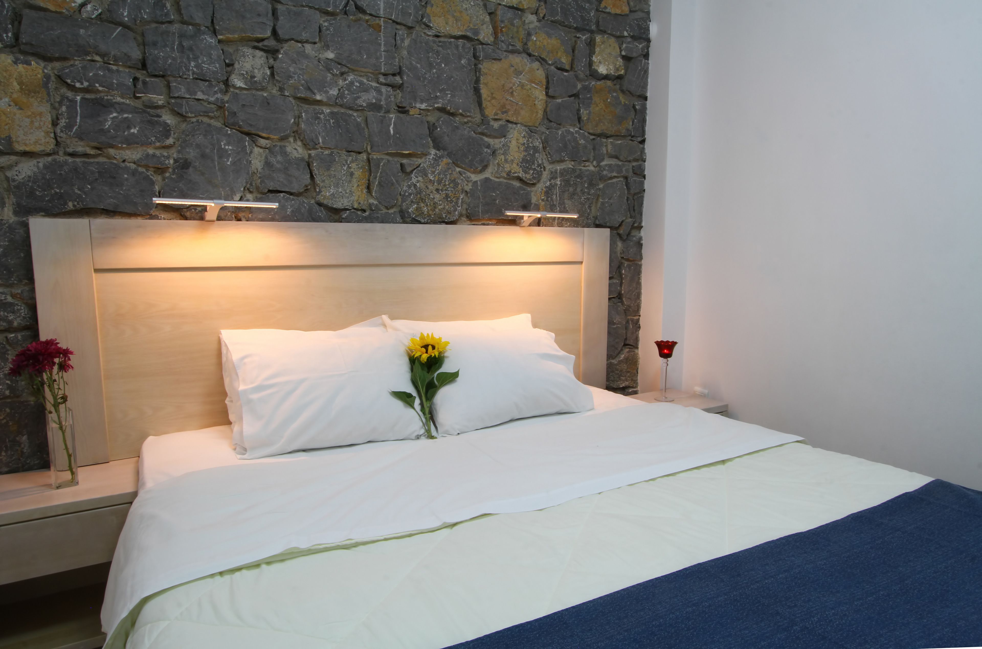 Una cama bien hecha con sábanas blancas, una flor amarilla y una vela roja sobre una mesita de noche.