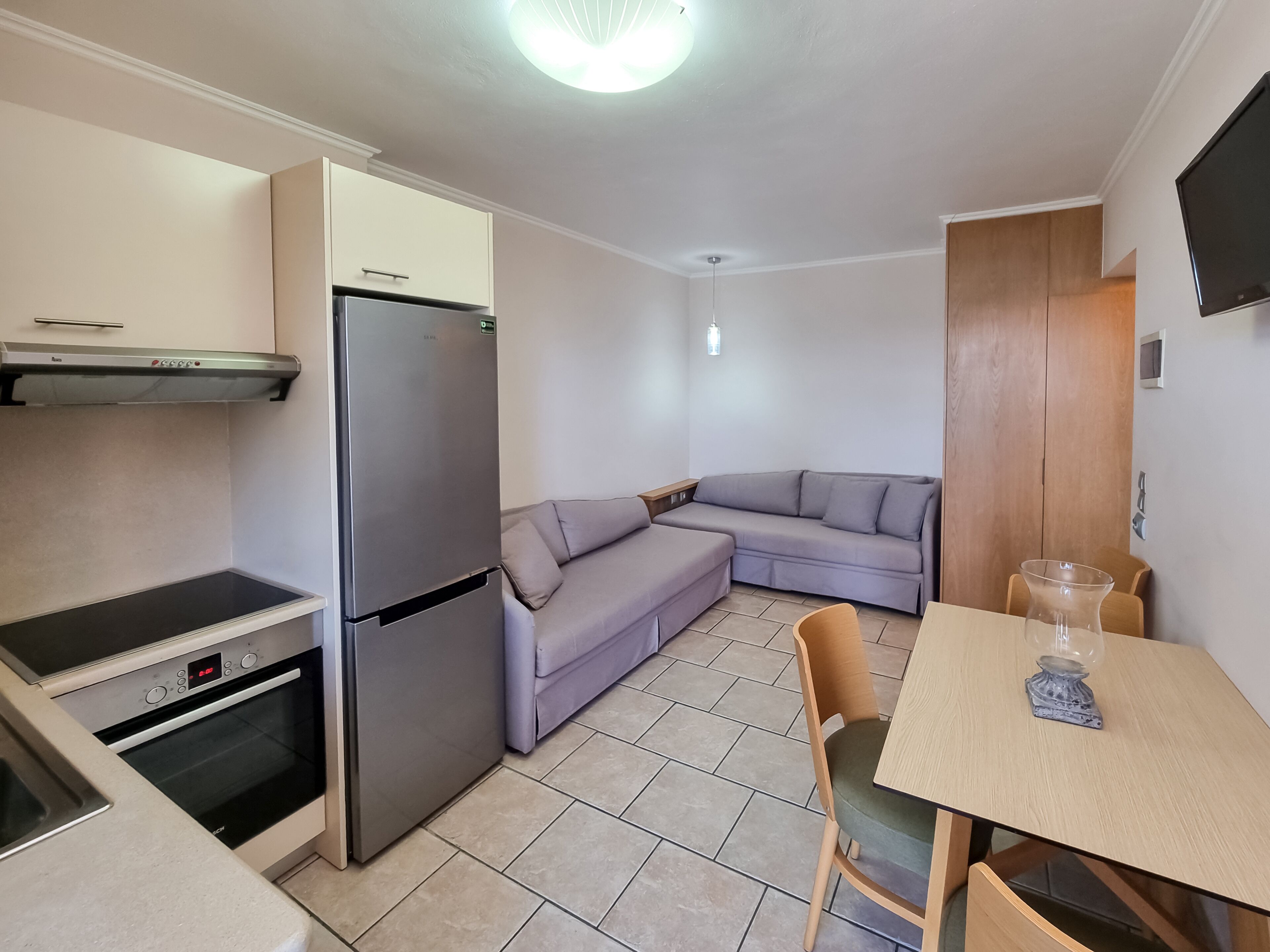 A compact kitchen with a refrigerator, oven, and a built-in stove. A gray sofa and a wooden dining table with chairs are in the living area.