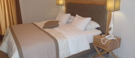 Una cama bien hecha con sábanas blancas y un edredón color beige, una mesita de noche con una lámpara y un teléfono, y un cabecero de madera.