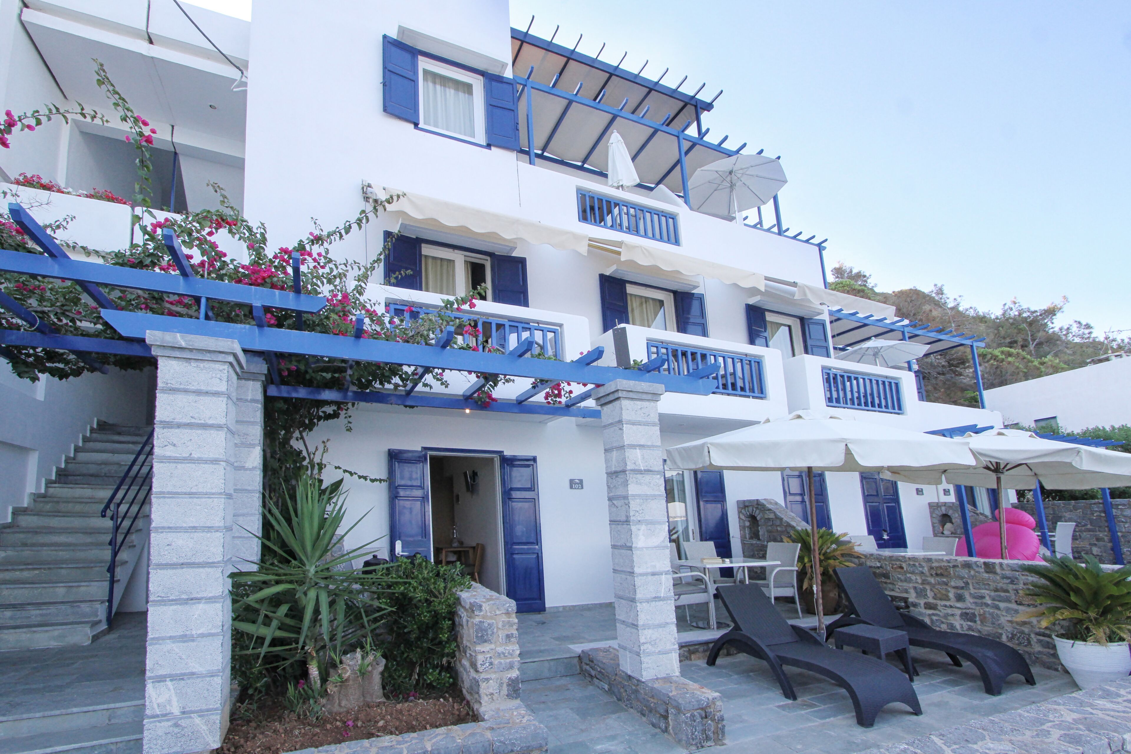 Un edificio de varios pisos con contraventanas azules, una escalera y mobiliario exterior con sombrillas.