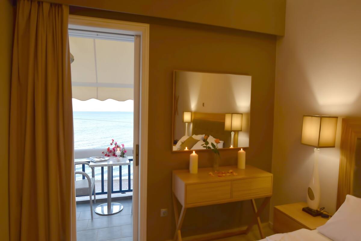 Habitación de hotel con balcón, una cama, una mesita de noche con lámpara y un espejo sobre el tocador.