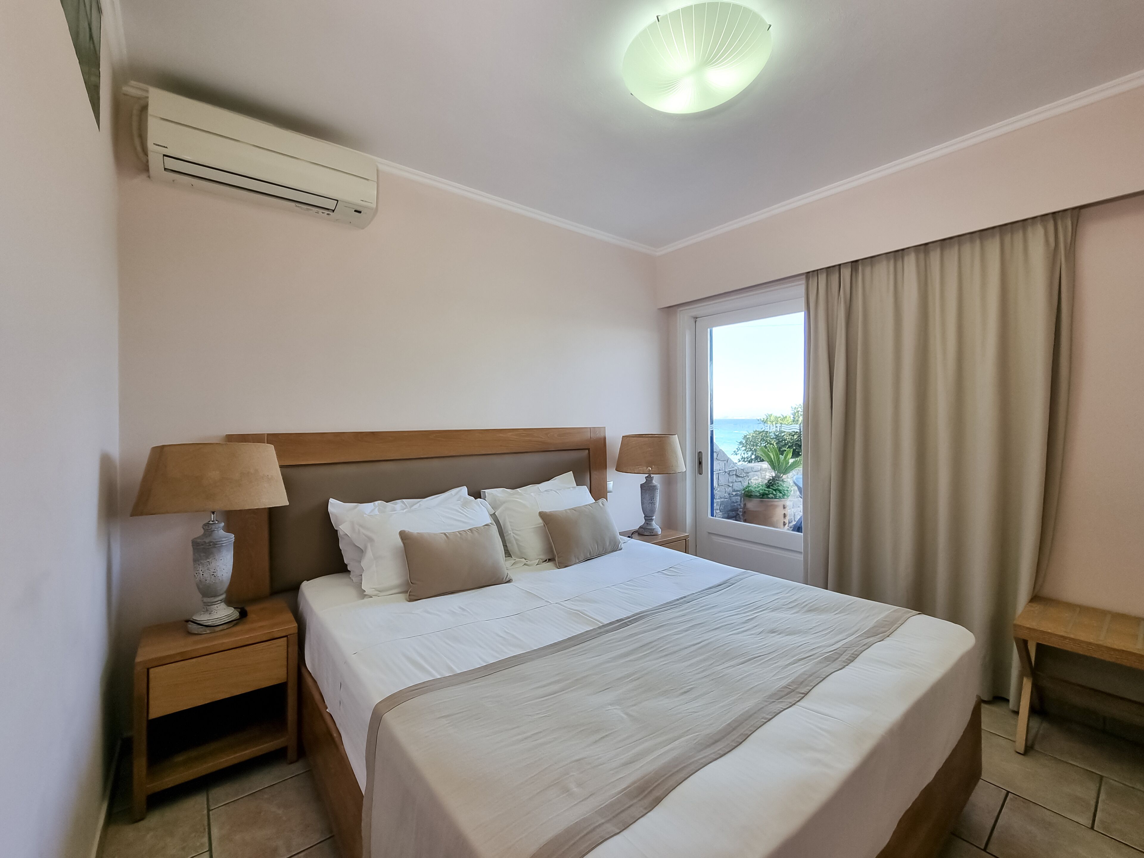 Une chambre d’hôtel avec un grand lit, deux tables de chevet avec des lampes, un luminaire au plafond, une fenêtre avec des rideaux et une vue sur la mer.