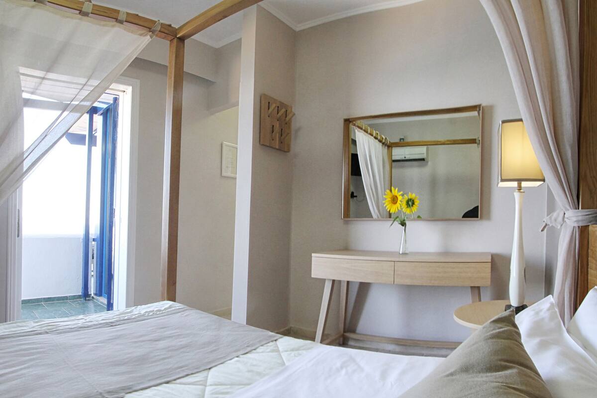 Un dormitorio con una cama, una mesita de noche con un florero de girasoles, un espejo y una lámpara.