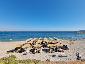 A beach with sun loungers and thatched umbrellas, a pebble beach, and the sea in the background. - Chios Xenia Studio & Apartments (Chios)