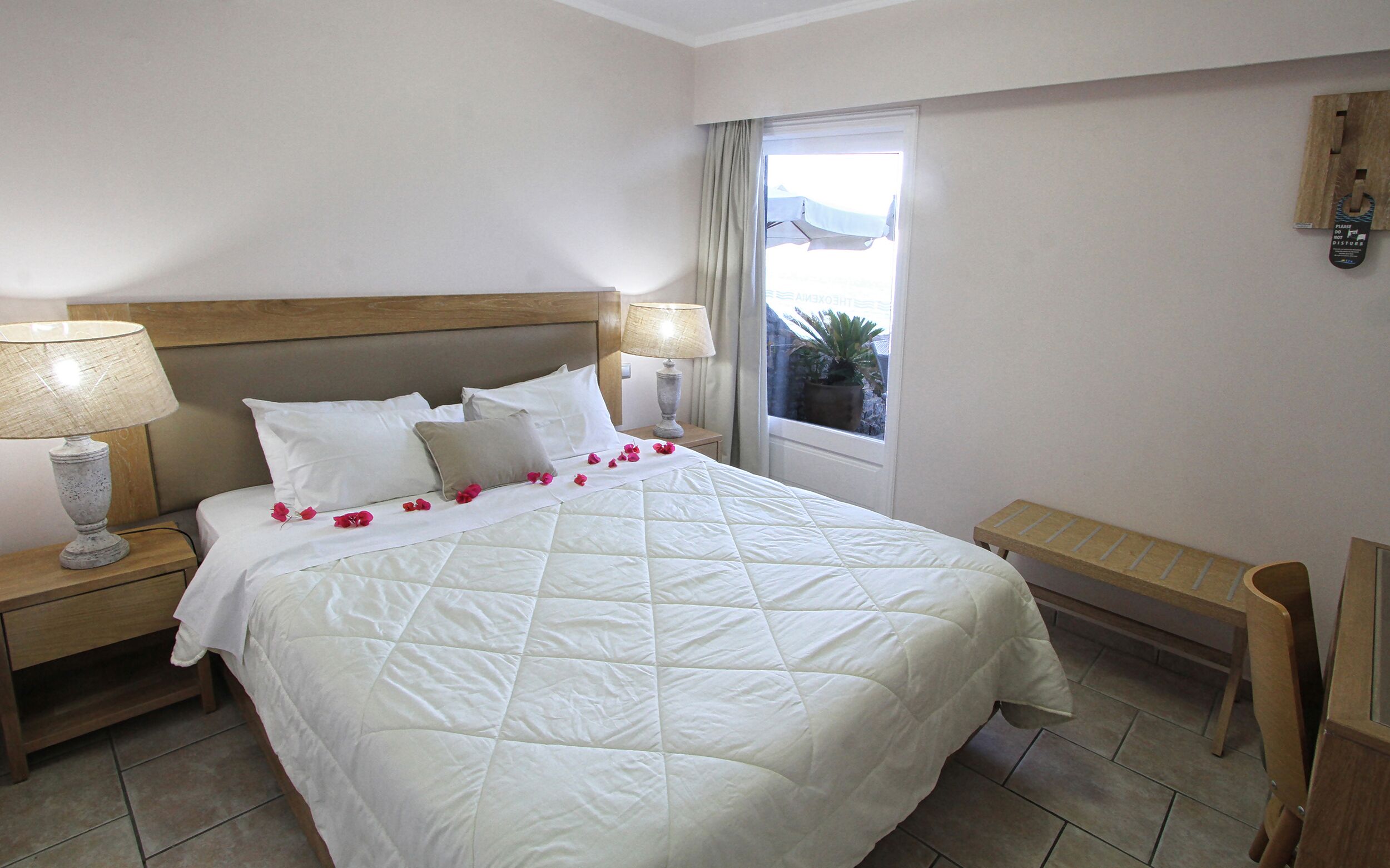 A neatly made bed with white linens and a quilted duvet, flanked by two bedside tables with lamps, a wooden bench, and a window with a view of the outdoors.