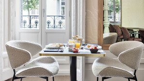 A breakfast table with a variety of food items and drinks, two upholstered chairs, and a view of the outside through a window with a balcony. - One Shot Recoletos (Madrid)