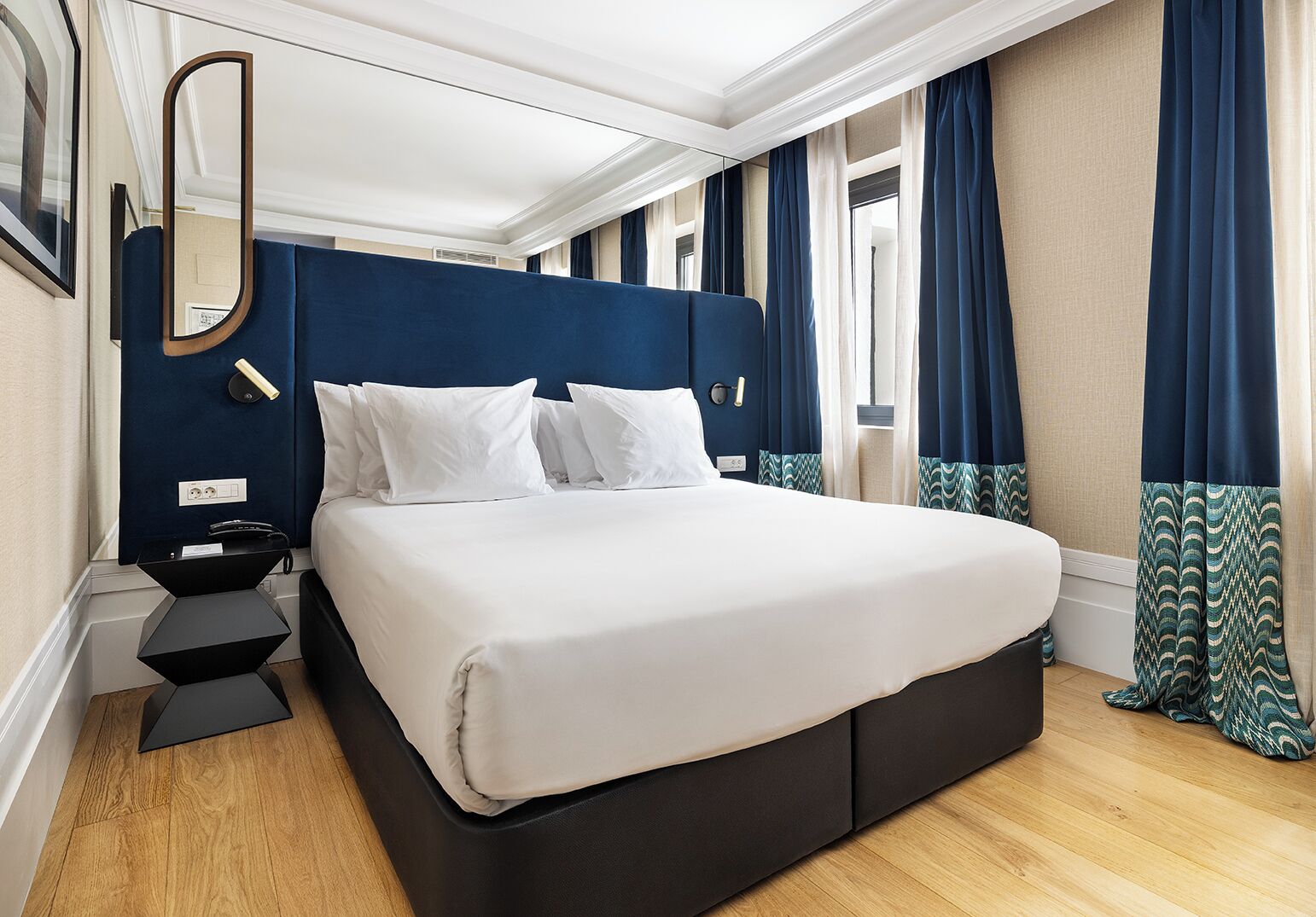 A modern hotel room with a large bed, dark blue headboard, white bedding, a bedside table with a phone, and a mirror.