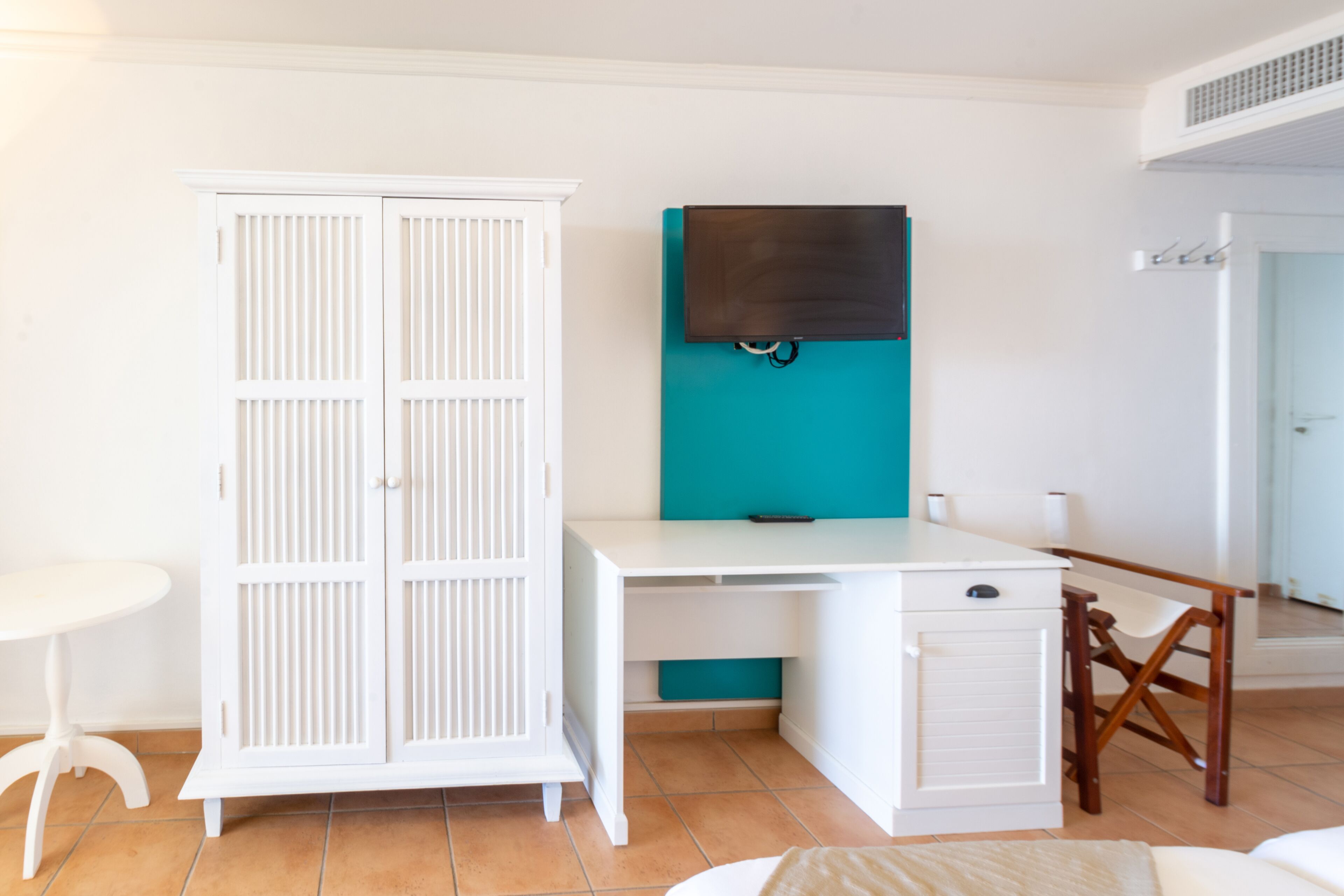 A room with a white wardrobe, a desk with a teal accent, and a small table.