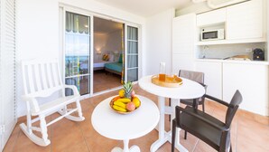 Un balcon avec un fauteuil à bascule blanc, une petite table sur laquelle est posé un panier de fruits, et un coin cuisine équipé d’un four à micro-ondes.