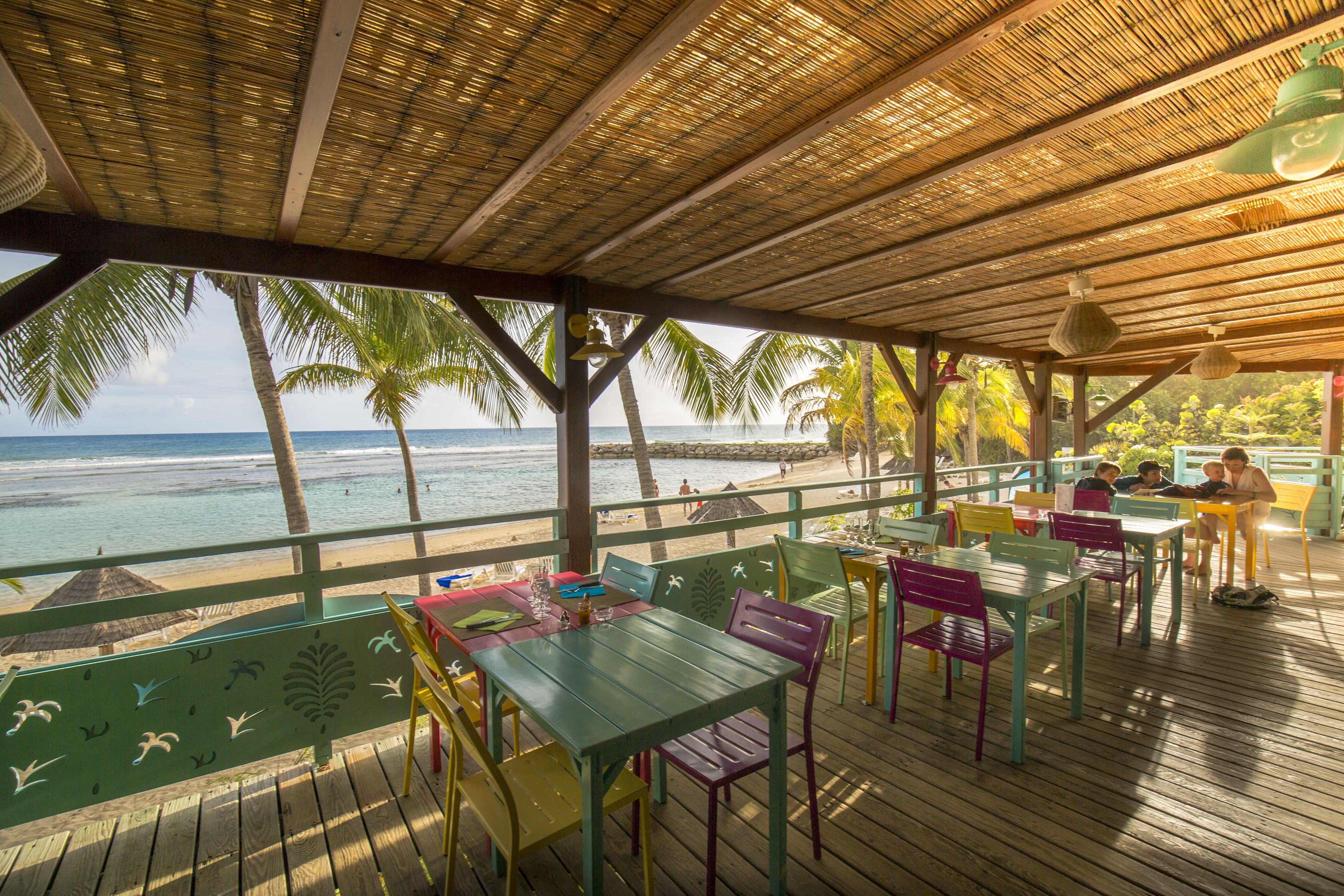 A covered outdoor seating area with colorful tables and chairs, overlooking a beach with palm trees and the ocean.