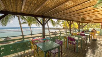 A covered outdoor seating area with colorful tables and chairs, overlooking a beach with palm trees and the ocean.