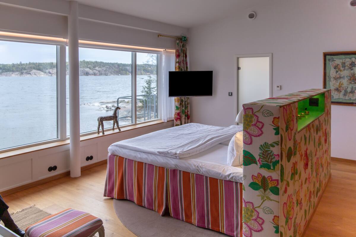 A bedroom with a large window overlooking a body of water, a bed with a floral-patterned headboard, a television mounted on the wall, and a small chair by the window.