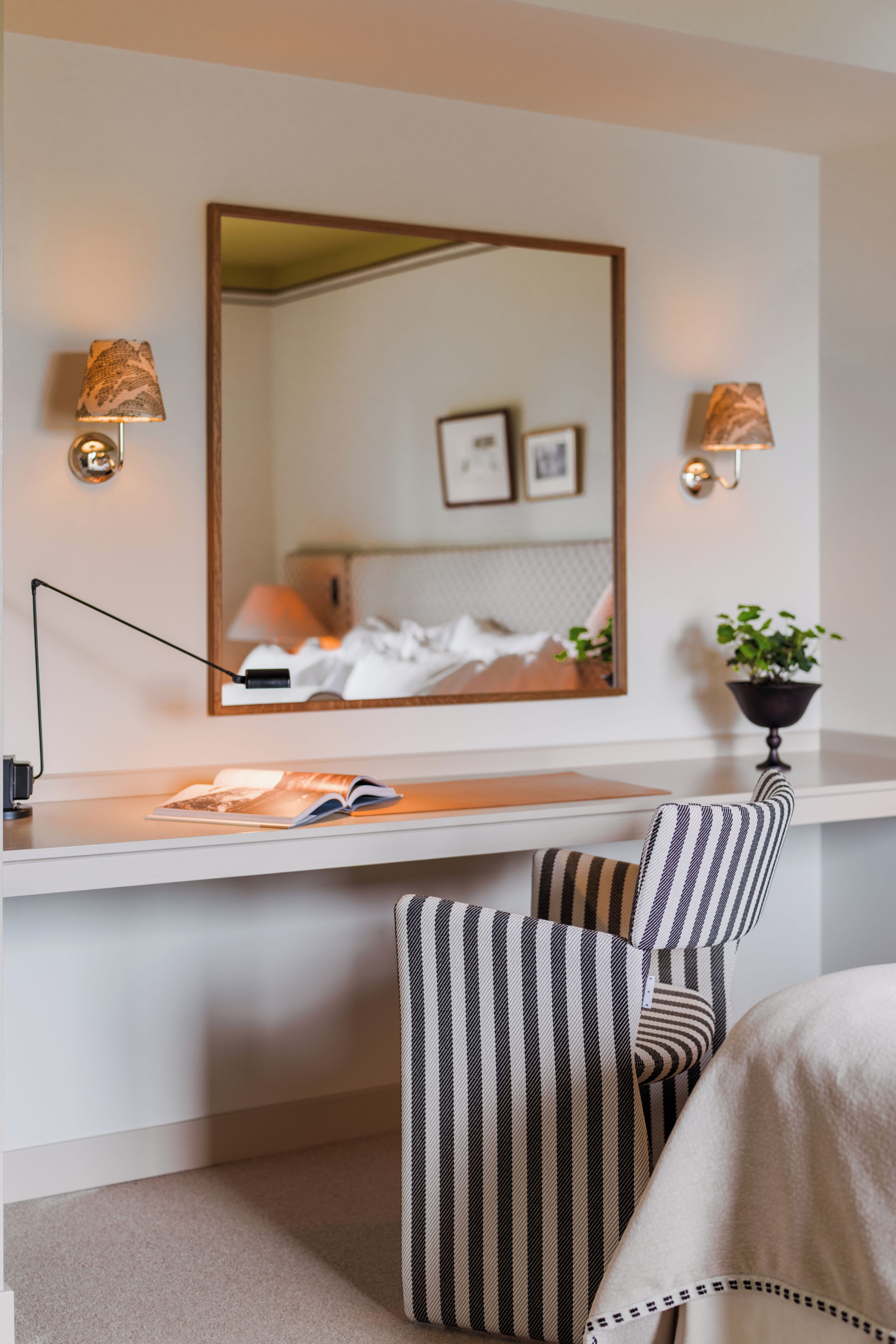 A hotel room with a desk, two lamps, a mirror, a bed, a plant, and a striped chair.