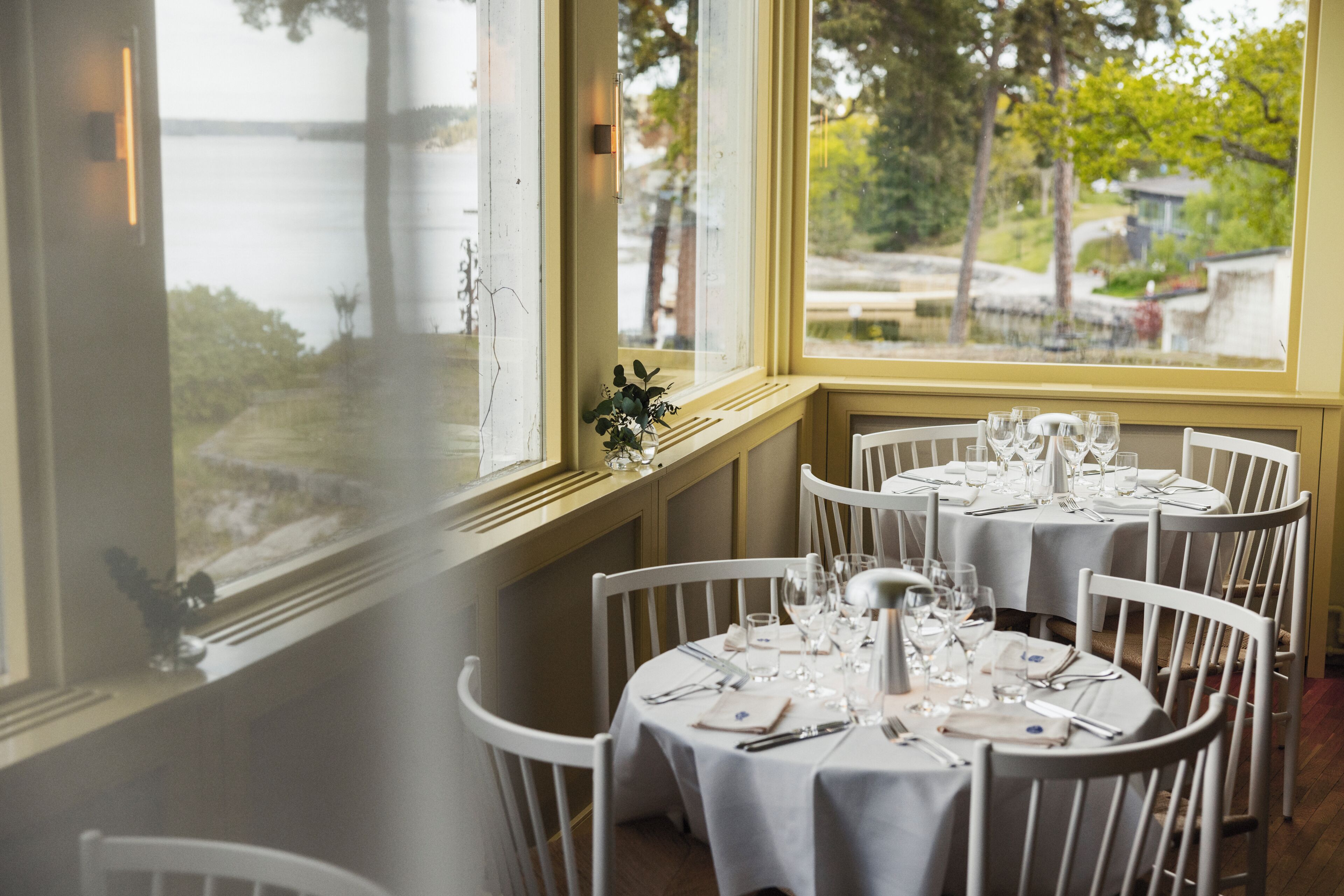 A dining area with round tables set for a meal, white chairs, and a view of a body of water through large windows.