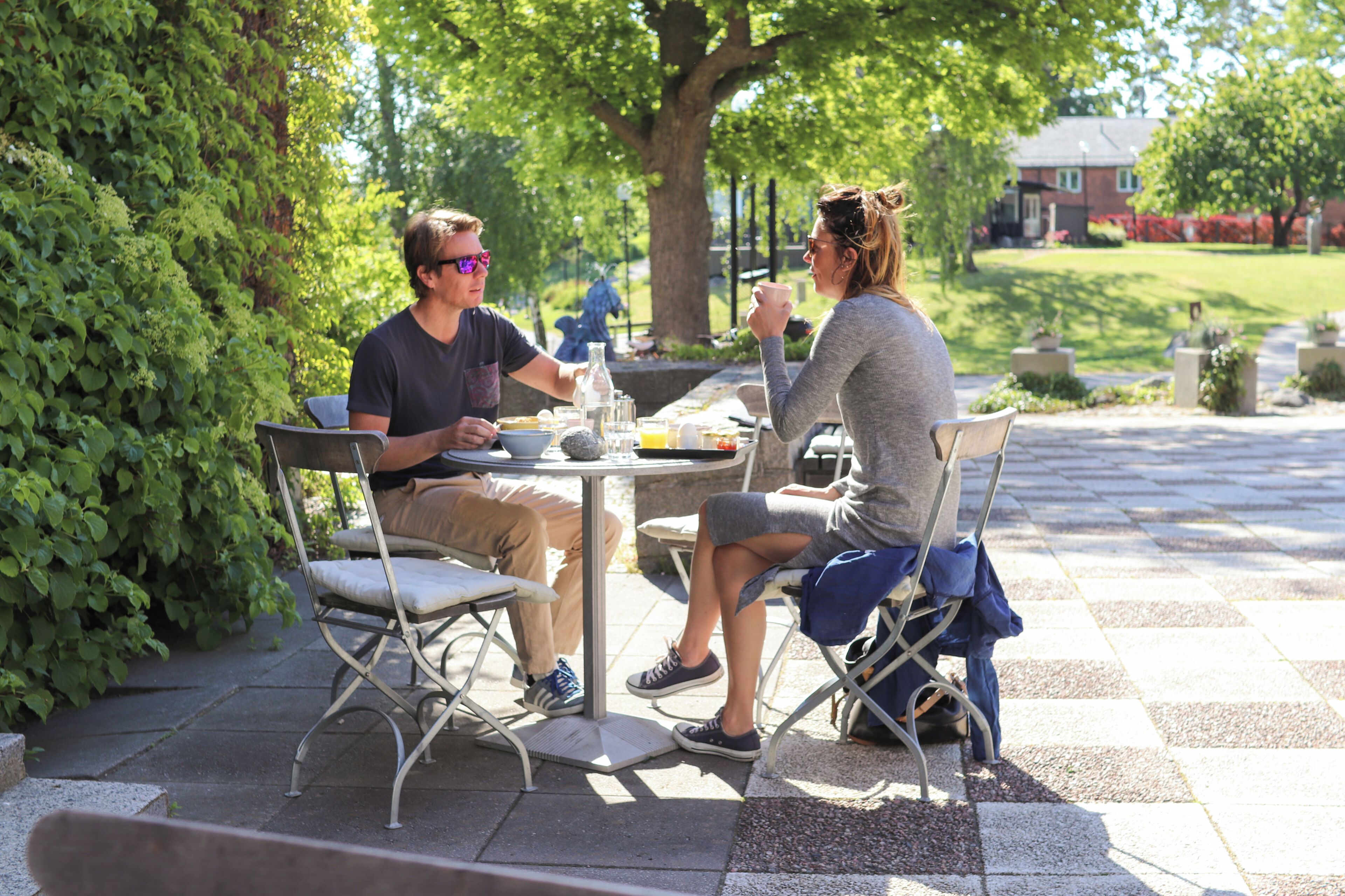 Two people dining at an outdoor table with chairs, surrounded by greenery and a paved area.