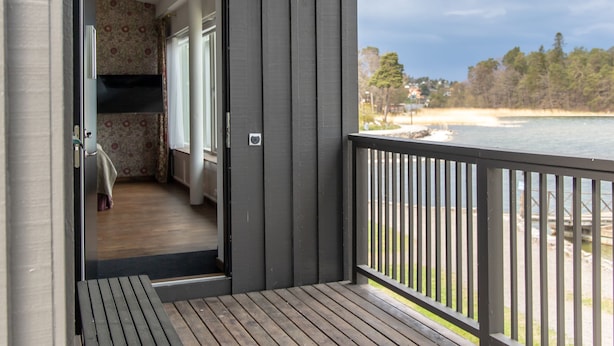 Una terraza de madera con un banco, una puerta que da a una habitación con papel tapiz floral, y vista a una playa y árboles.