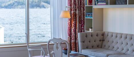 A room with a large window overlooking a body of water, a sofa, a chair, a small table, and a bookshelf.