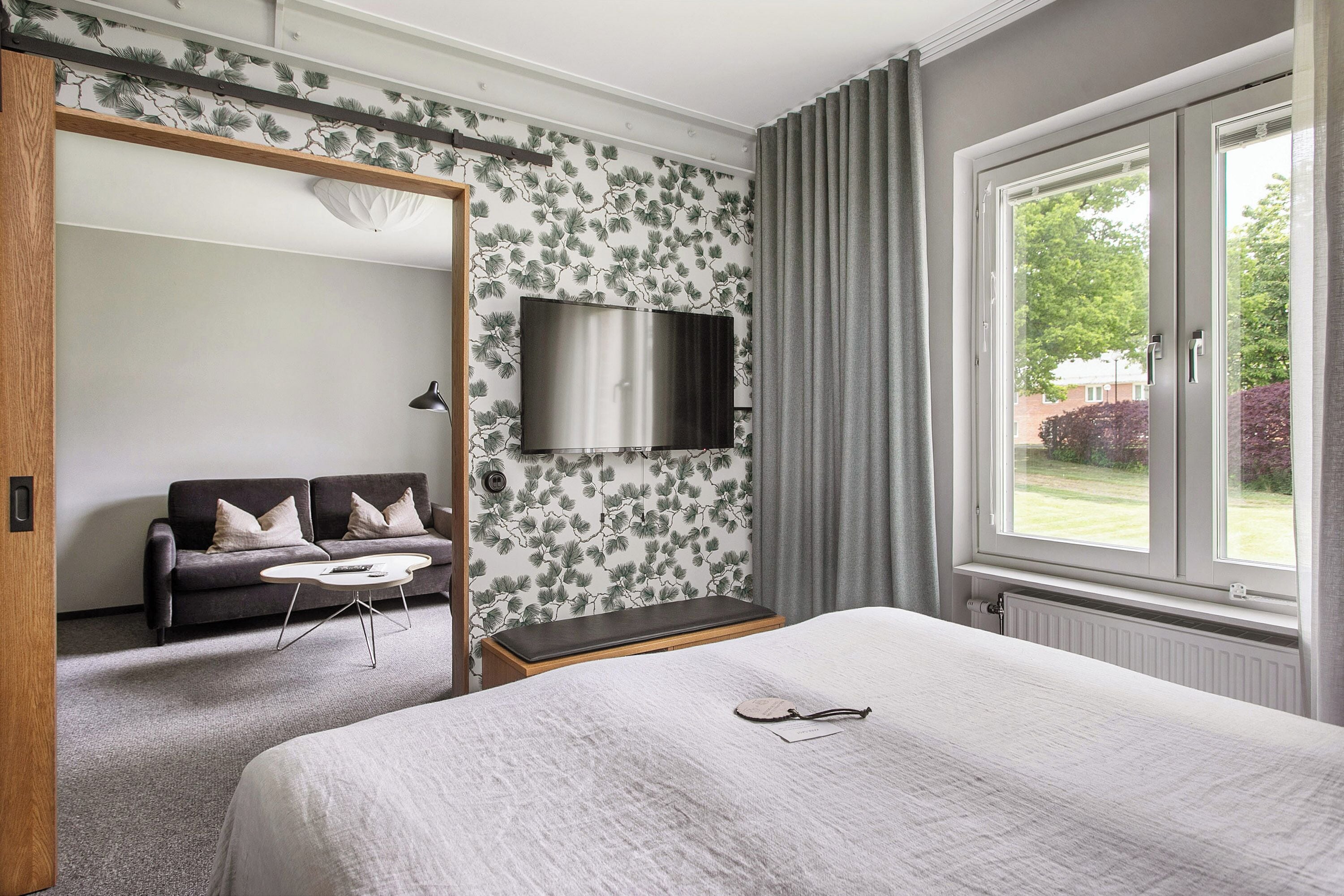 A bedroom with a bed, a TV mounted on the wall, a sofa, and a window with curtains.