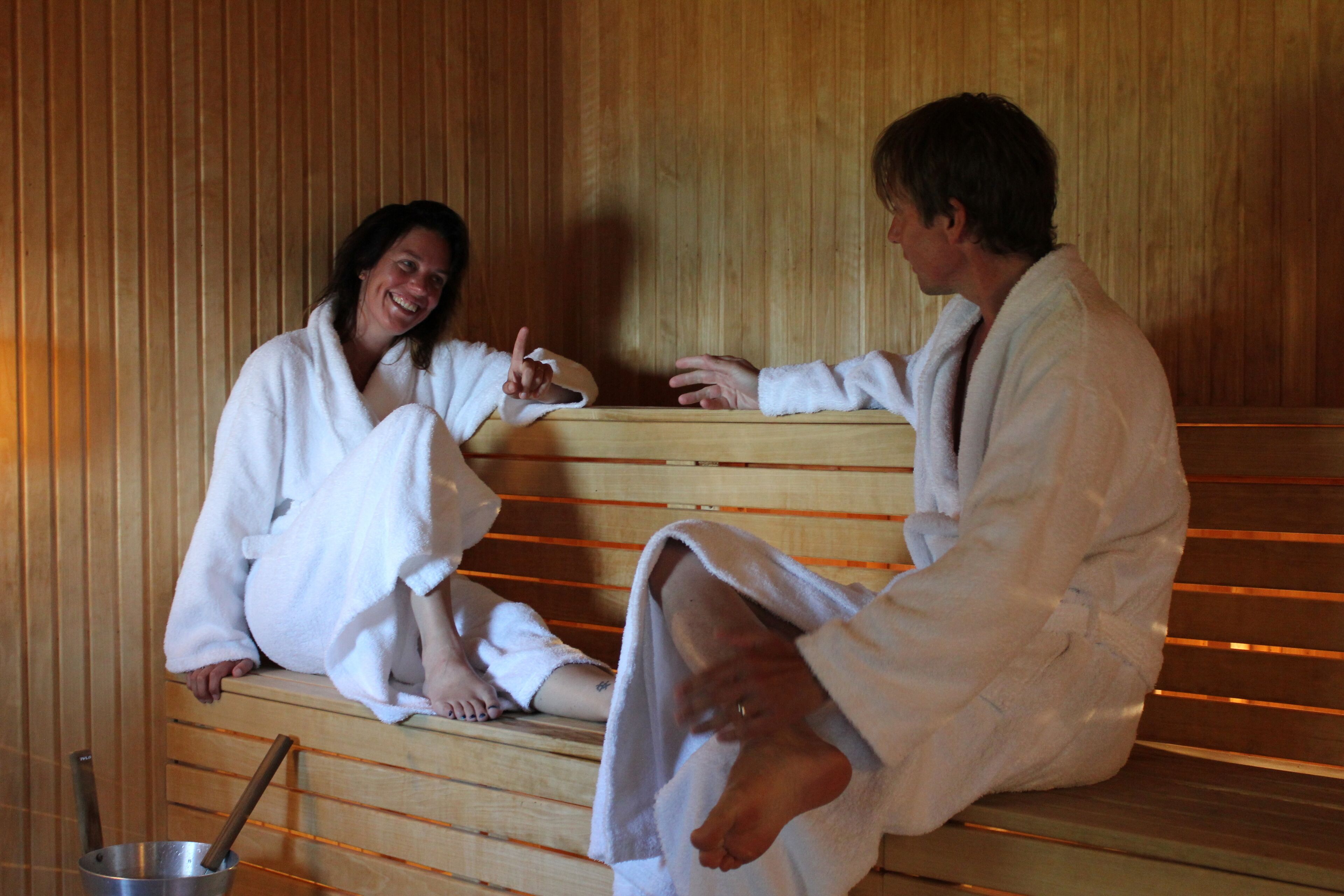 Duas pessoas com roupões brancos sentadas em bancos de madeira em uma sauna.