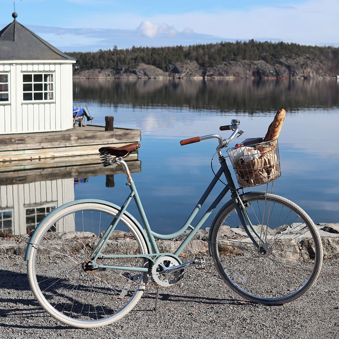 A bicycle with a basket and a bag, parked near a dock by a calm lake with a wooden structure in the background.
