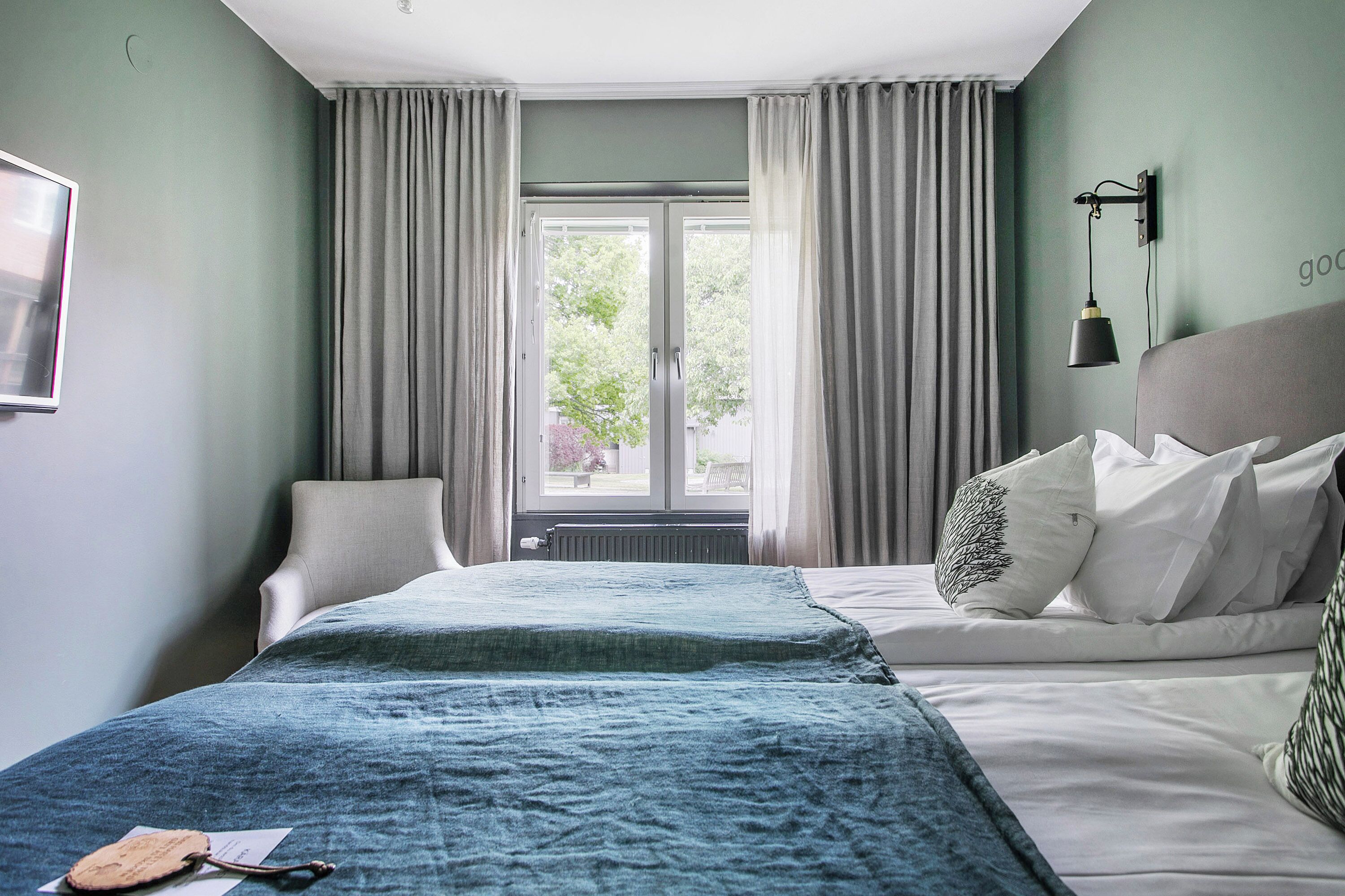 A hotel room with a bed, a chair, a window with curtains, and a wall-mounted lamp.