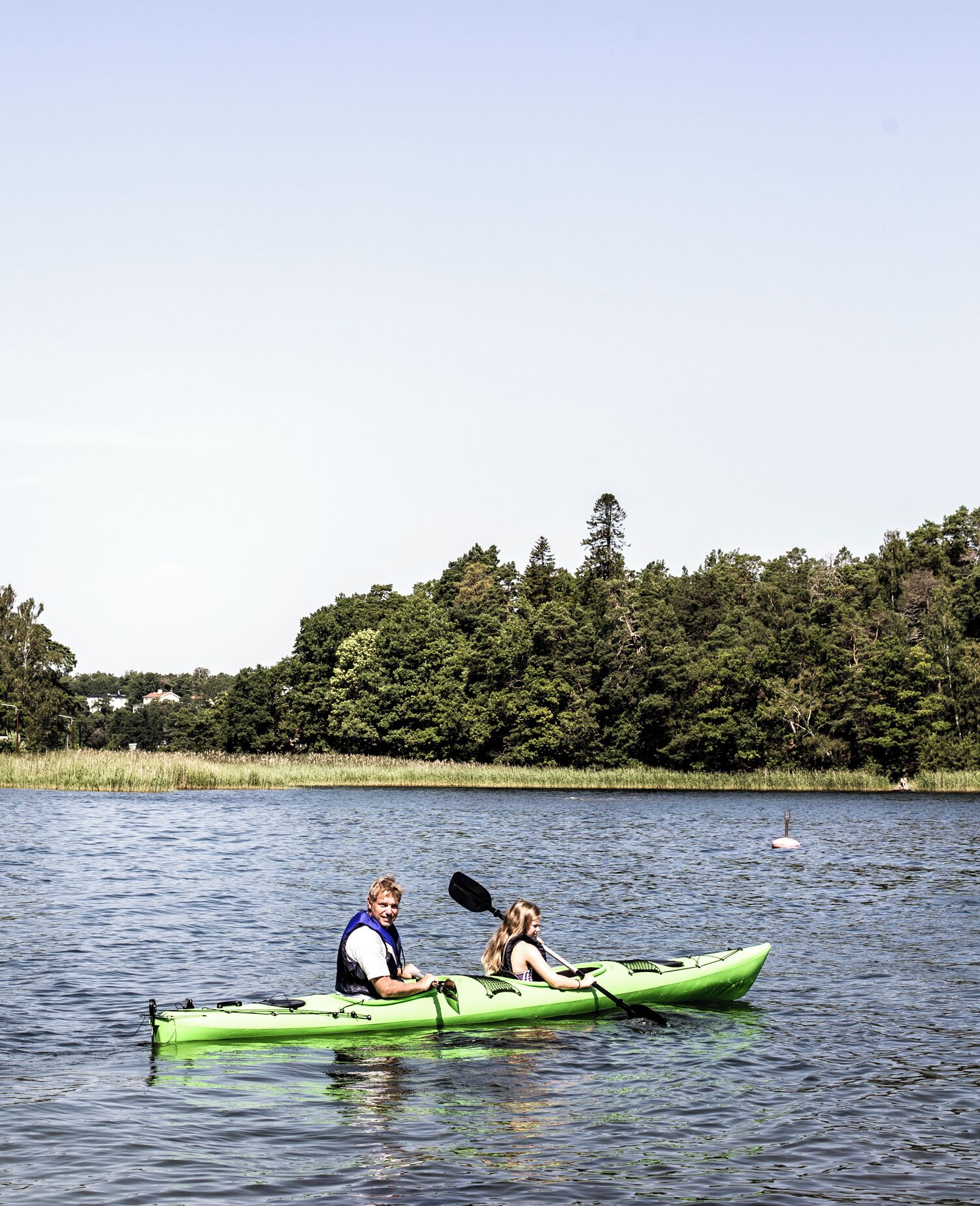 Two people in a green kayak on a calm lake with a forested shoreline.