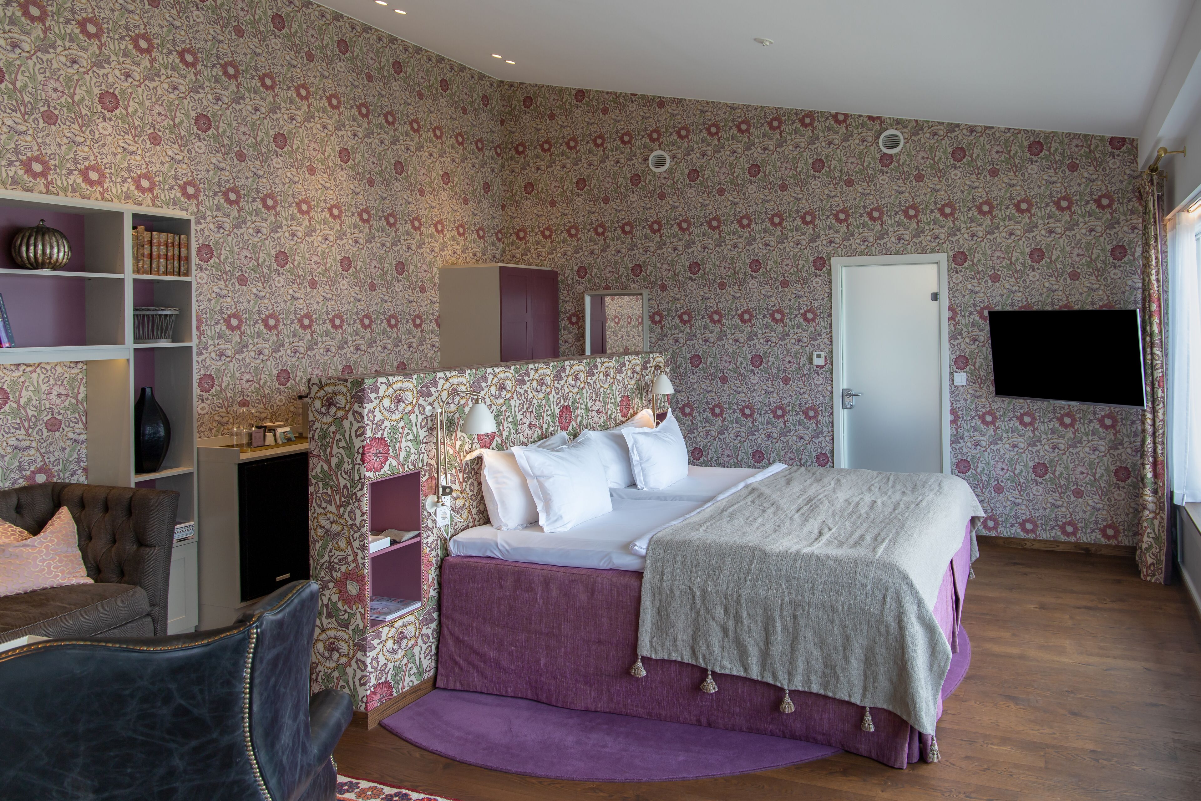 A bedroom with a floral wallpaper, a bed with a purple bedspread, a chest of drawers, a desk, and a television on the wall.