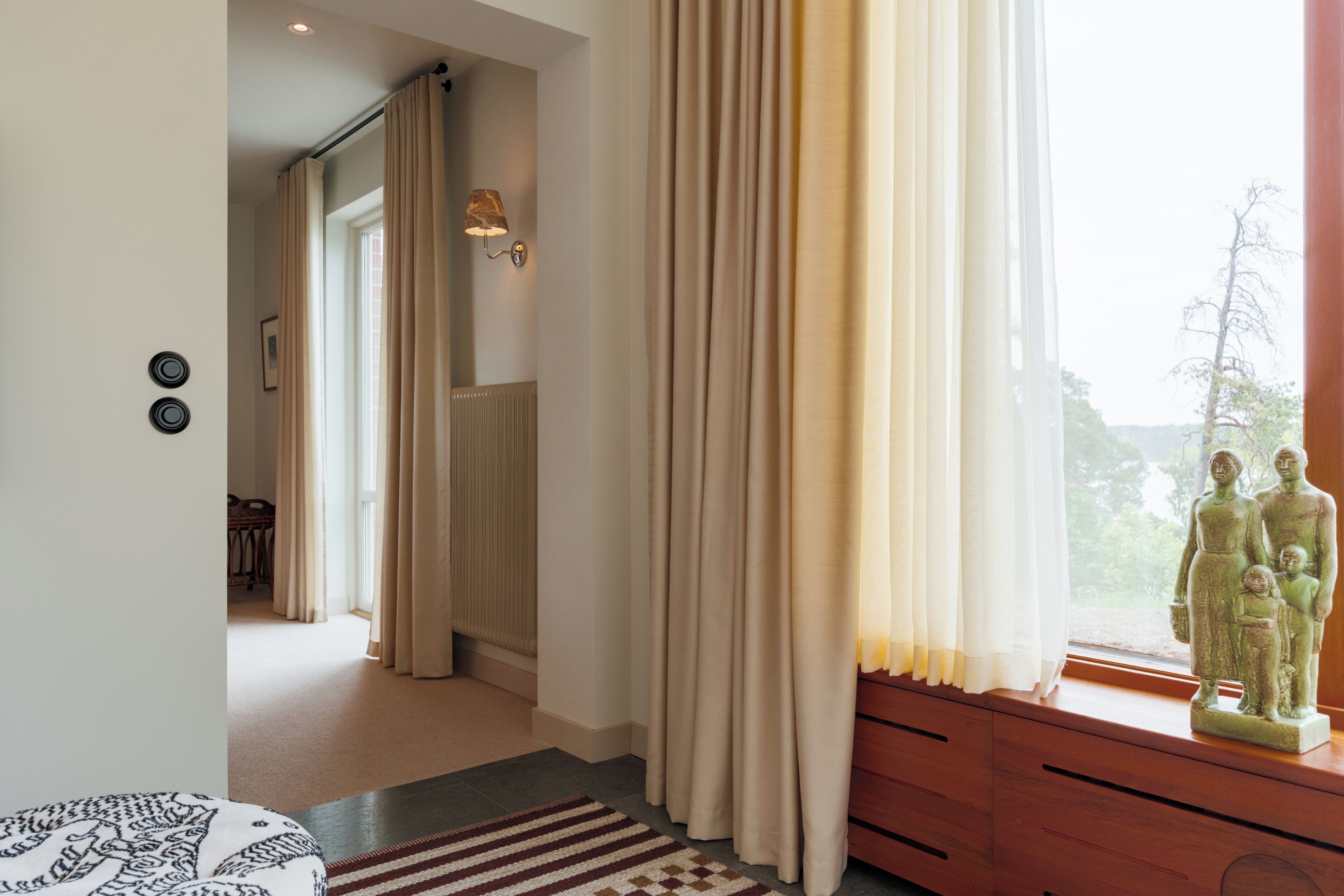 Une pièce avec une fenêtre, des rideaux, une statue, un radiateur et un tapis à motifs.