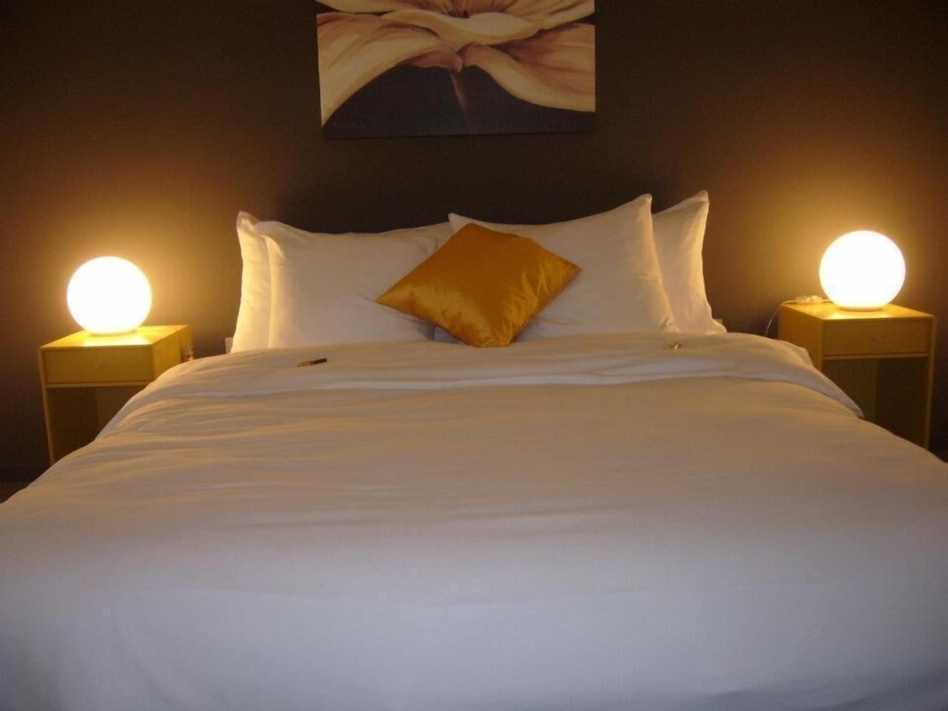 A neatly made bed with white linens and a single gold pillow, flanked by two bedside lamps on matching nightstands.