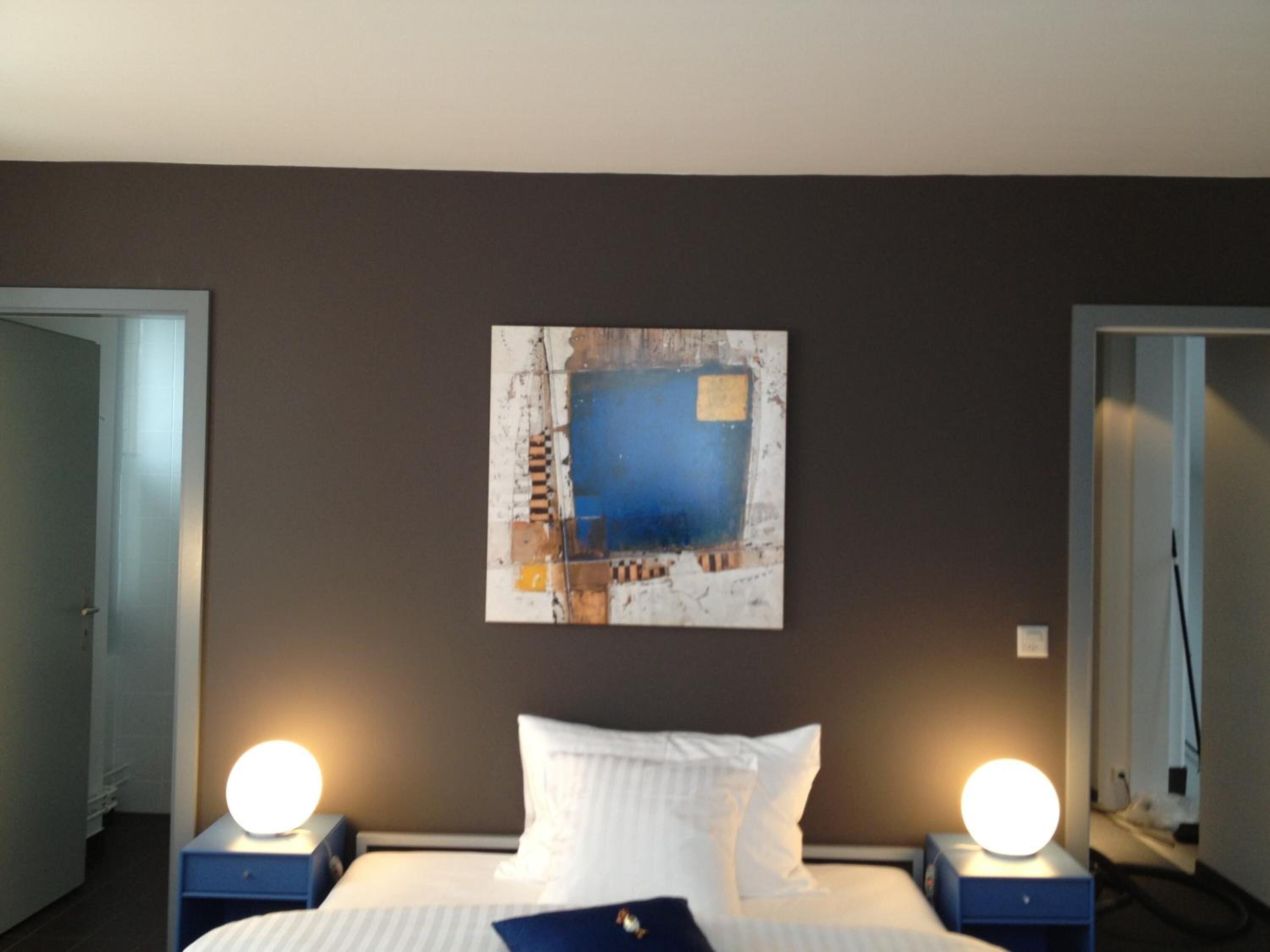 Une chambre d’hôtel avec un lit, deux tables de chevet avec des lampes, un tableau abstrait accroché au mur et une porte sur le côté gauche.