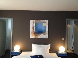 A hotel room with a bed, two bedside tables with lamps, a wall-mounted abstract painting, and a door to the left. - Hôtel au Boeuf Rouge (Crassier)