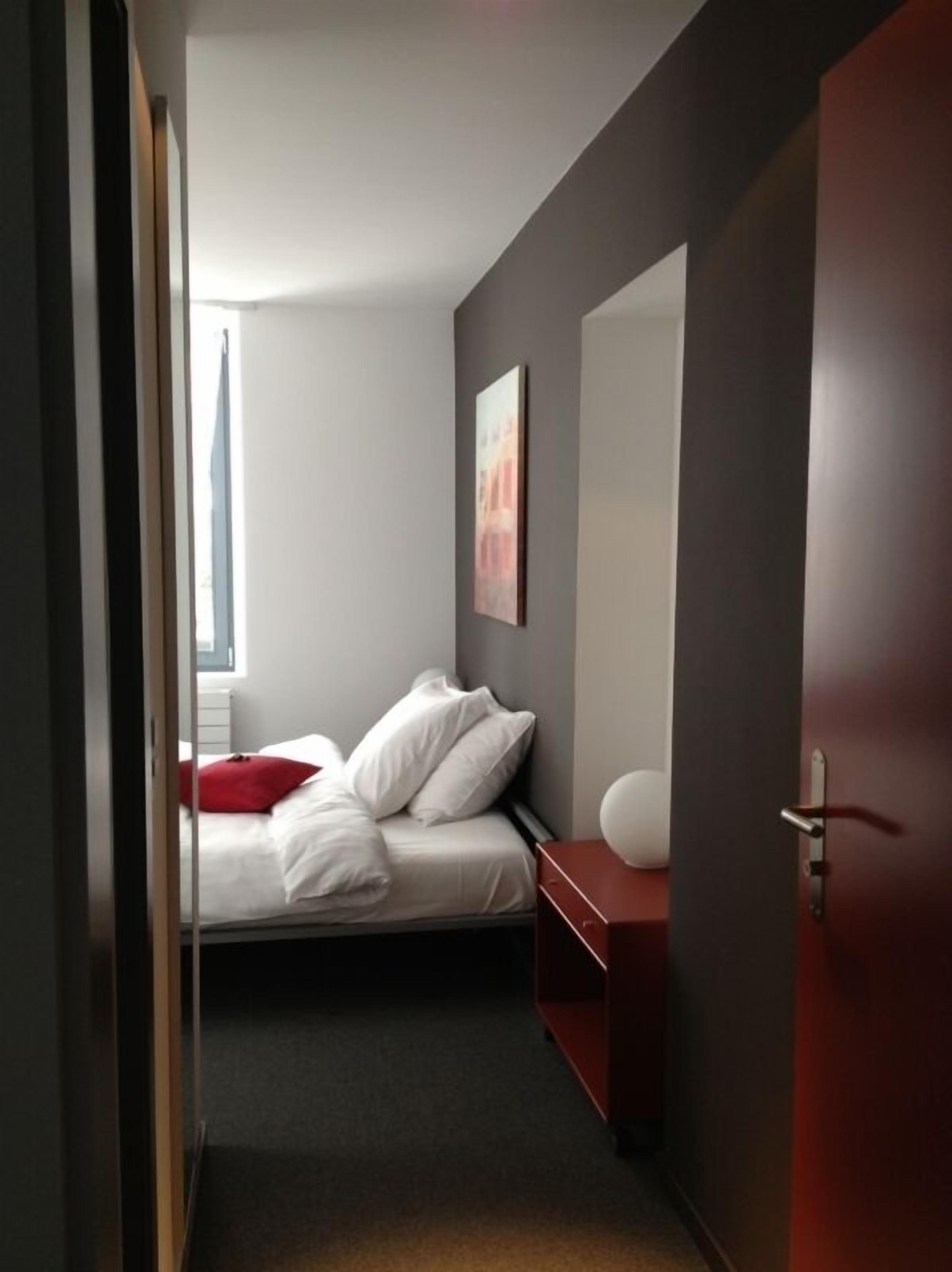 Una habitación de hotel moderna con una cama, una mesita de noche, una mesita de noche roja, un espejo grande y un cuadro en la pared.