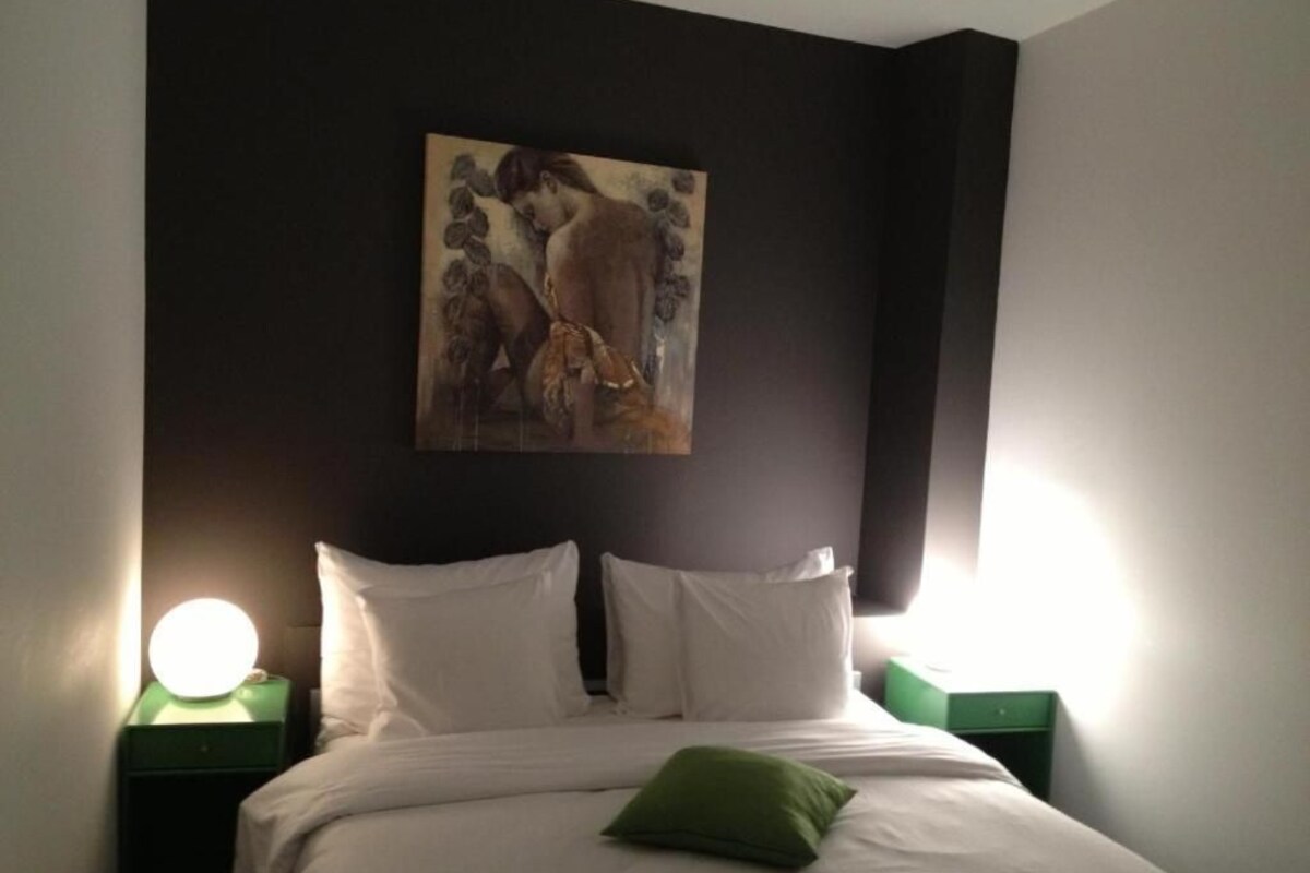 A hotel room with a bed, white pillows, a green pillow, a painting on the wall, and two bedside lamps.