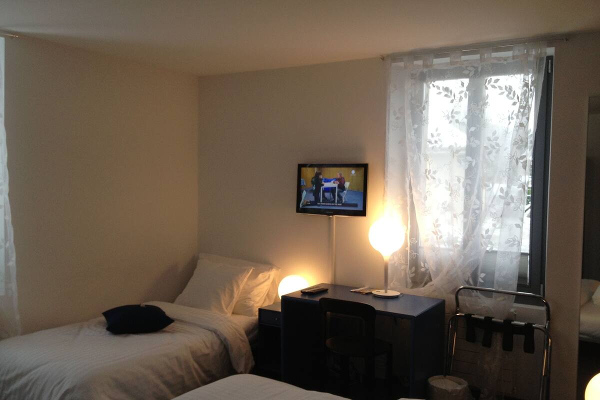 A hotel room with two beds, a desk, a television mounted on the wall, and a window with sheer curtains.