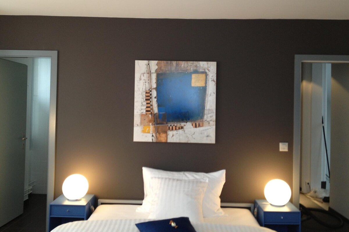A hotel room with a bed, two bedside tables with lamps, a wall-mounted artwork, and a door to the left.