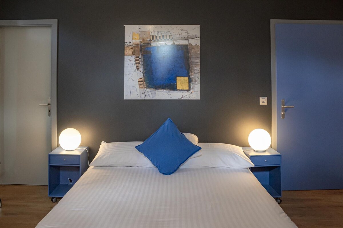 A hotel room with a bed, two bedside tables with lamps, a door, and a wall-mounted artwork.