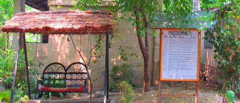 A rustic wooden structure with a thatched roof and a bench, a bulletin board with a program, and a potted plant.