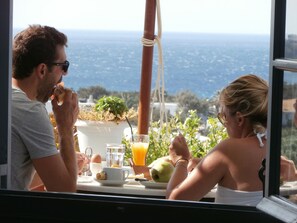 A dining table with breakfast items, a potted plant, and a view of the sea.
