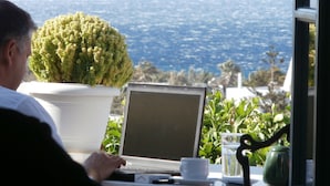 A laptop on a table with a potted plant, a cup, and a glass of water, overlooking a coastal view. - Galini Bungalows (Tinos)