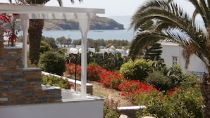 A coastal view with a white building, red flowers, and palm trees. - Galini Bungalows (Tinos)