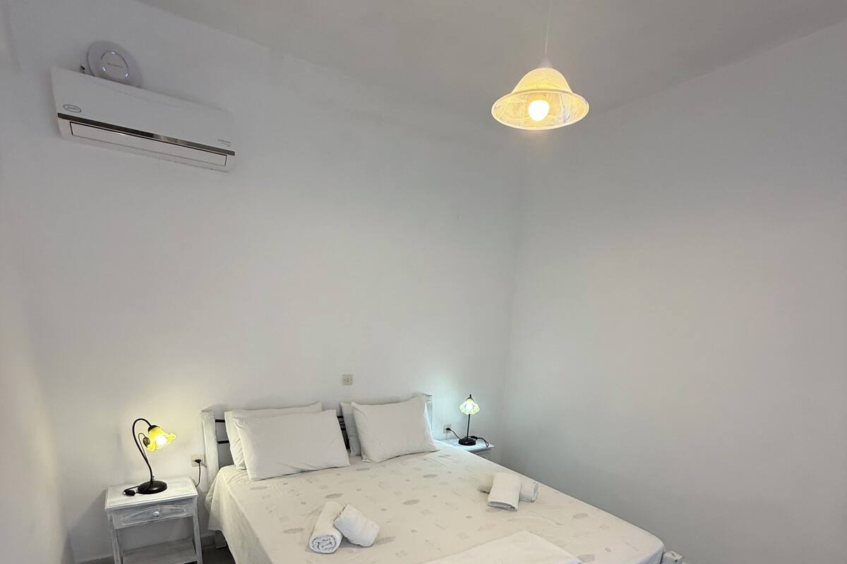 A minimalist bedroom with a bed, bedside lamps, a wall clock, and an air conditioning unit.
