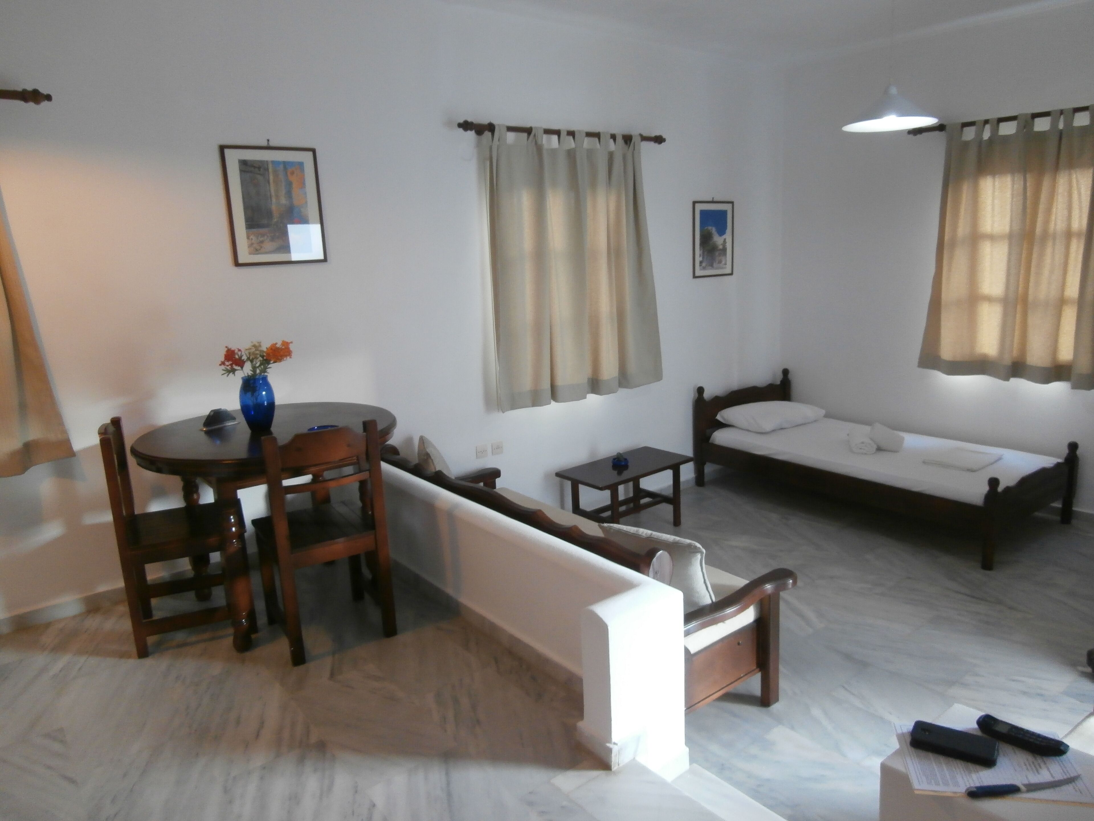 A room with a bed, a sofa, a dining table with chairs, a small table, and a vase with flowers.
