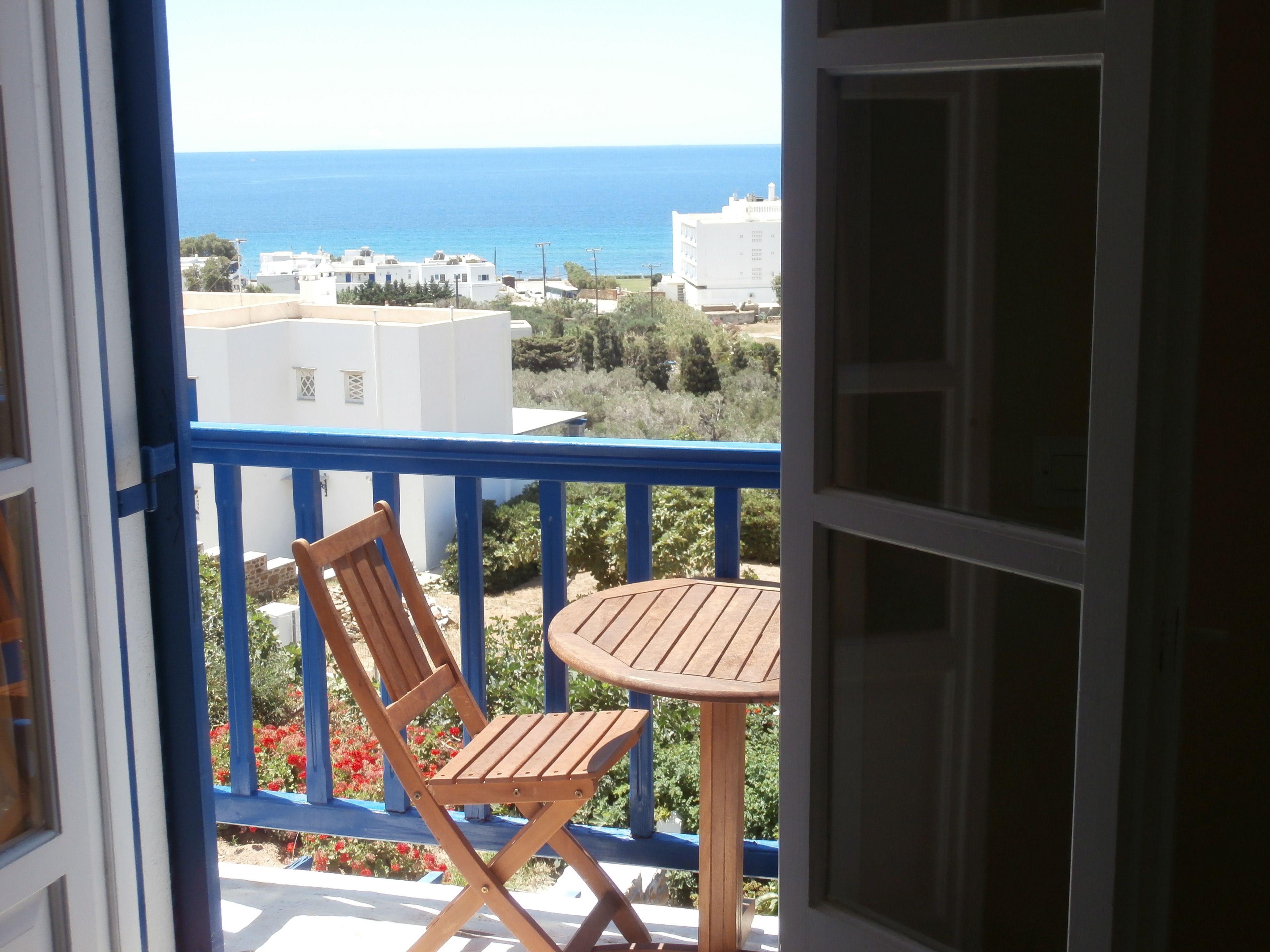 A balcony with a wooden chair and table, overlooking a coastal area with buildings and the sea.