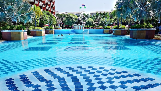 Une grande piscine extérieure, dont le fond est recouvert de carreaux formant un motif, entourée de palmiers et bordée d’un bâtiment doté de balcons.