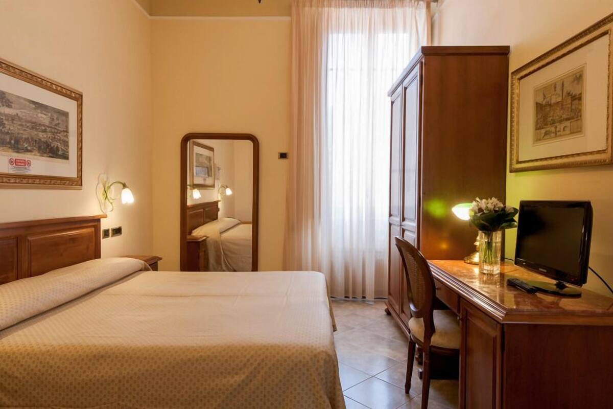 Habitación de hotel con cama, escritorio con computadora, espejo y ventana con cortinas.