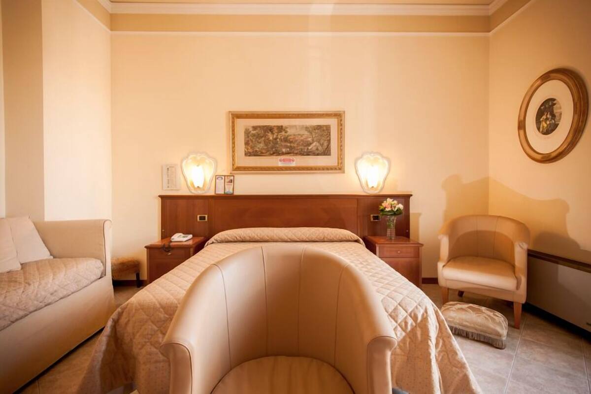 Habitación de hotel con una cama grande, una mesita de noche con una lámpara, una silla y un cuadro en la pared.