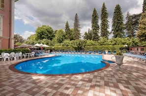 Piscina con sillas de descanso y sombrillas, rodeada de árboles y un edificio.