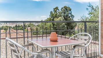 Un balcón con mesa y sillas, una planta en maceta y vistas a árboles y edificios.